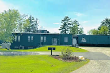 Slate gray industrial building set on a meadow with trees behind.